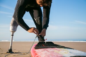 ウェットスーツと義肢を身に着け、砂の上でサーフボードを足首に結びつける男性サーファー