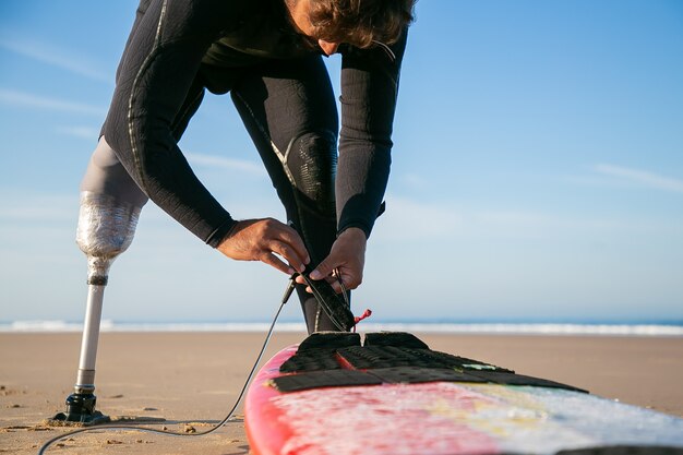 Мужчина-серфер в гидрокостюме и протезе, привязывающий доску для серфинга к лодыжке на песке