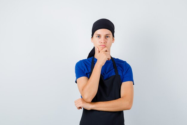 Male teen cook keeping hand on chin in t-shirt, apron and looking pensive. front view.