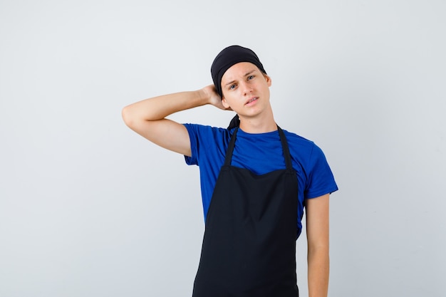 남자 10대 요리사는 티셔츠, 앞치마를 입고 머리 뒤로 손을 잡고 사려깊은 앞모습을 보고 있습니다.