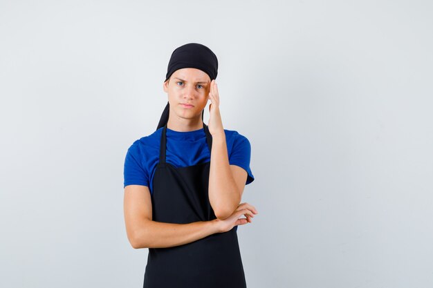 Male teen cook keeping hand on temple in t-shirt, apron and looking serious. front view.