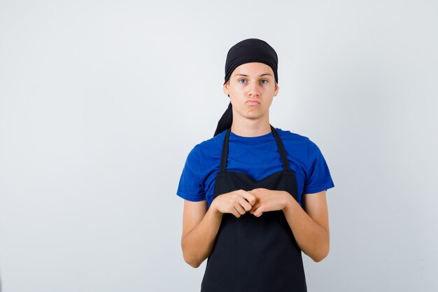 男性の10代の料理人は、Tシャツ、エプロンで考えているポーズで立って、思慮深く見えます。正面図。