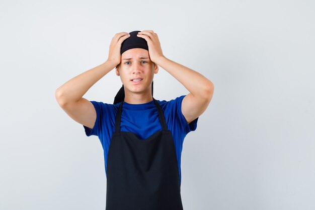 Мужчина-подросток готовит в футболке, фартуке с руками на голове и выглядит раскаявшимся, вид спереди.