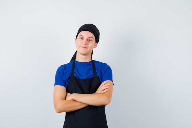 Male teen cook with hands crossed in t-shirt, apron and looking pleased. front view.