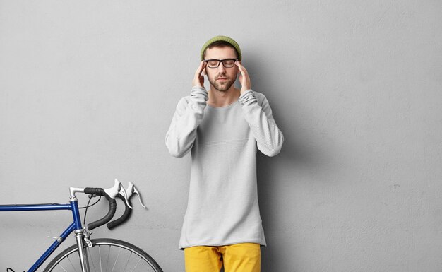 Male teenager wearing sweater and colorful trousers, trying to concentrate while standing in his room near his modern bicycle. Attentive cyclist trying to solve all obstacles on his way