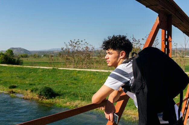 Male tourist on a bridge over the river holding cellphone and carrying backpack