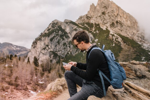 Male tourist with short dark hair resting on stone and texting message after climbing to mountains