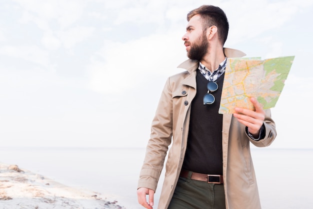 Free photo male traveler standing on beach holding map in hand looking away