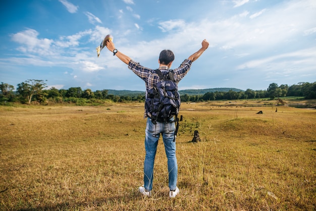 A male traveler with a backpack carrying a map and standing on the meadow.