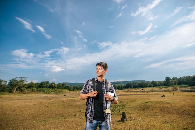 A male traveler with a backpack carrying a map and standing on the meadow.