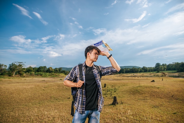 A male traveler with a backpack carrying a map and standing on the meadow.