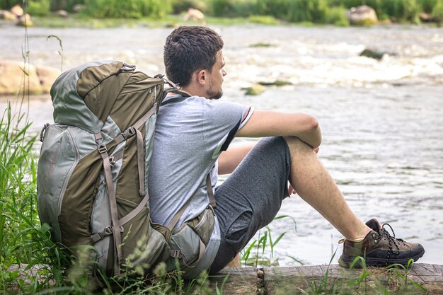 大きなハイキング用バックパックを背負った男性旅行者が川の近くで休んでいます。