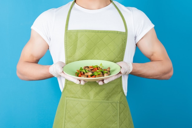 Male waiter in green apron holding delicious meal on blue wall