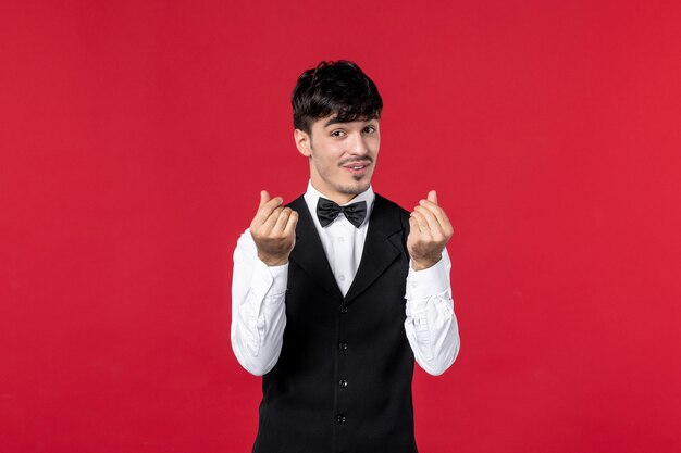 male waiter in a uniform with butterfly making money gesture on neck on red background