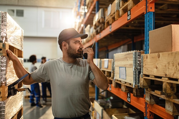 Male warehouse worker communicating with someone over walkietalkie while standing among shelfs in distribution warehouse
