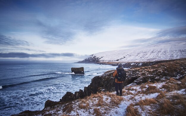 Male wearing a backpack and a jacket standing on the snowy hill while taking a picture of the sea