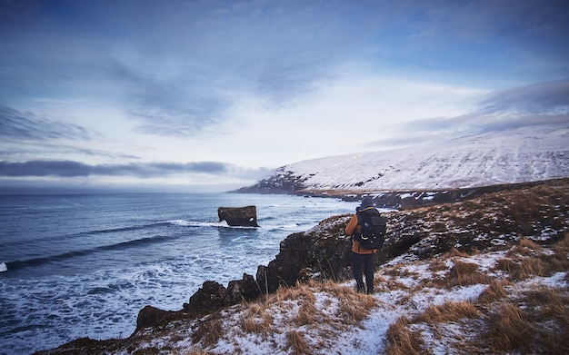 海の写真を撮りながら雪に覆われた丘の上に立っているバックパックとジャケットを着ている男性