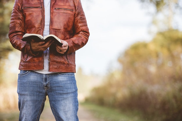Мужчина в кожаной куртке стоит на пустой дороге и читает библию с размытым пространством