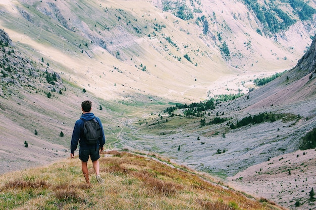 後ろから山のショットに囲まれた景色を楽しみながら崖の上に立っているバックパックを持つ男性