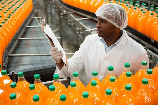 Male worker reading clipboard while inspecting bottles in juice factory