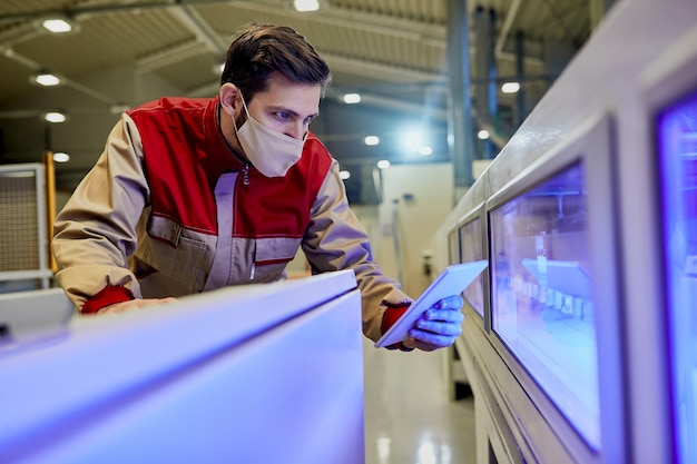 Male worker with face mask using touchpad while supervising woodworking production line machine