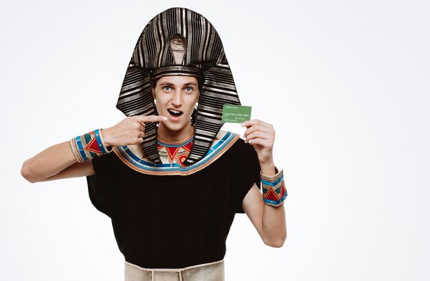Человек в древнем египетском костюме держит кредитную карту, указывая на нее указательным пальцем, весело улыбаясь, счастлив и доволен на белом