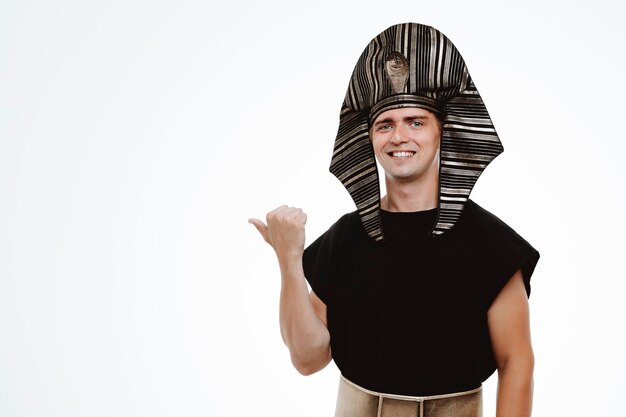 Man in ancient egyptian costume smiling confident pointing with thumb to the side on white