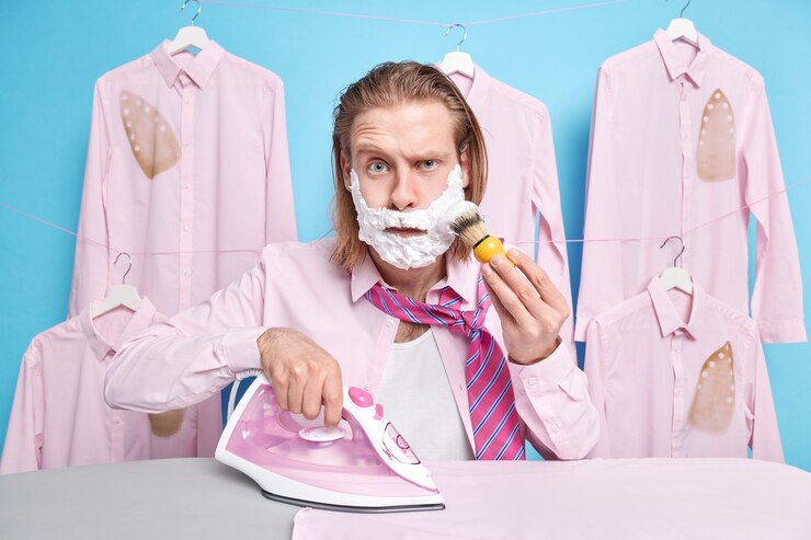 Free Photo man applies shaving gel on cheeks and raises eyebrows wears shirt and tie irons clothes prepares for party being at home