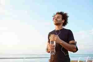 Free photo man athlete drinking water out of plastic bottle after hard running workout. dark-skinned male sportsman looking at the sky while running, enjoying view