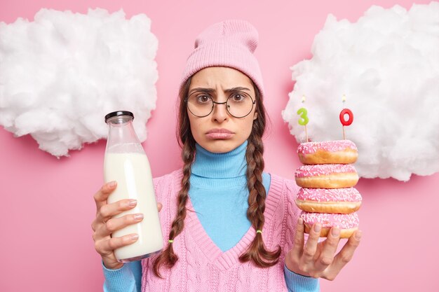 man in bad mood celebrates her 30th anniversary alone holds doughnuts with number birthday candles bottle of milk wears hat round spectacles poses indoor on rosy 