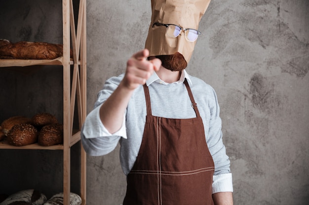 Free photo man baker standing with paper bag on head pointing.