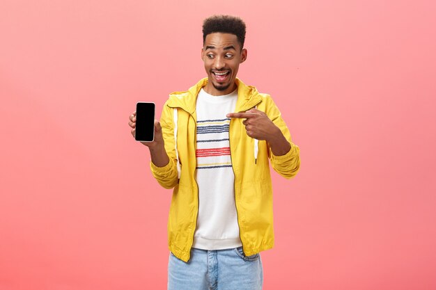 Man being amazed with cool new phone cannot hide happiness from device purchase holding smartphone pointing at gadget screen with excited and impressed face posing over pink background