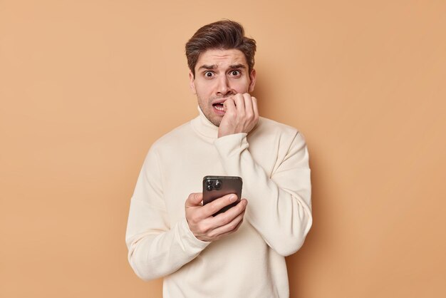 man bites finger nails reads chat nervously holds mobile phone wears casual jumper has scared expression made huge mistake isolated on beige. What should I do help