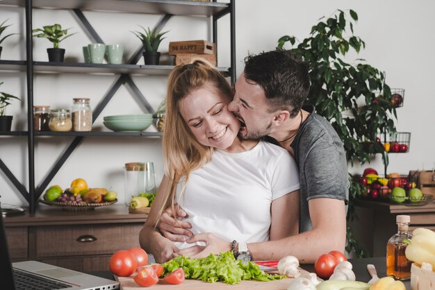 Man biting on woman cheeks standing behind the kitchen counter with vegetables