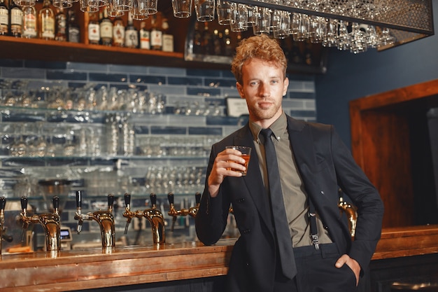 Man in a black suit drinks alcohol. Attractive guy drinks whiskey from a glass..