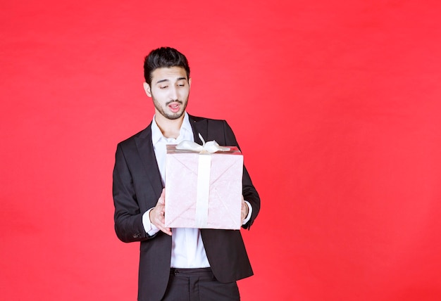 Man in black suit holding a purple gift box and looks confused and thoughtful.