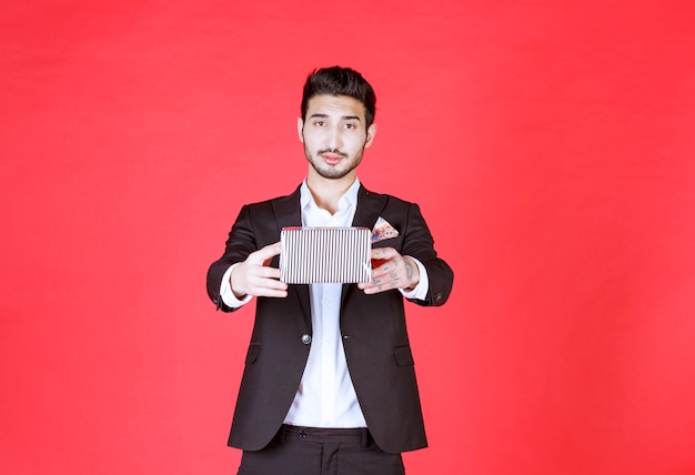 Man in black suit holding a silver gift box and pointing the person he would present it.