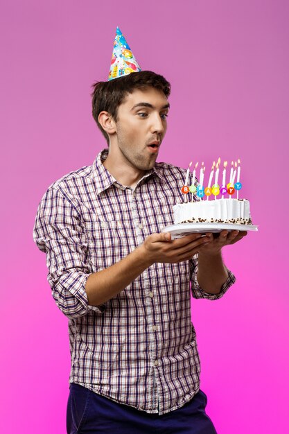 Man blowing out candles on birthday cake over purple wall.