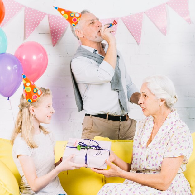 Man blowing party horn while girl giving birthday gift to her grandmother