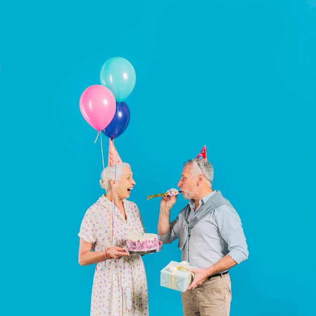 青い背景に誕生日のケーキを抱いている彼の妻の間にパーティーホーンを吹く男