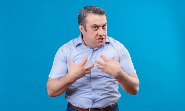 A man in blue shirt emotionally shows aggression and anger with hands pointing at himself on a blue space