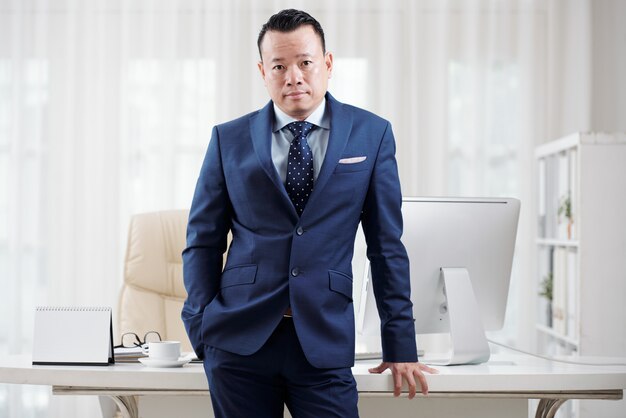 Man in blue suit leaning on his office table to pose for a picture