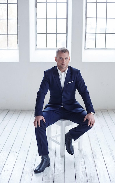 A man in blue suit sitting in white room with windows on background.