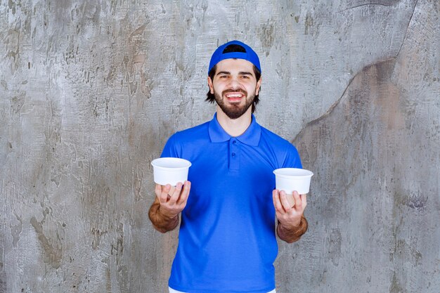 파란색 유니폼을 입은 남자가 양손에 두 개의 테이크아웃 플라스틱 컵을 들고 있습니다.