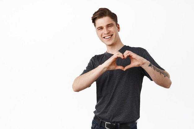  man, boyfriend shows I love you heart sign and smiles cute at you, laughing while confessing his feelings, standing on white