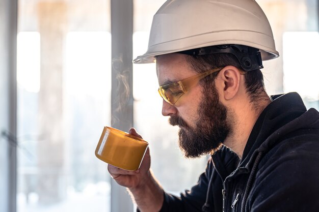 A man builder in a helmet and goggles drinks a hot drink