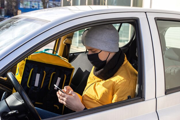 A man in the car with black medical mask is on his phone, backpack on the seat