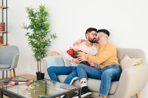 Man carrying baby in hand kissing his boyfriend sitting on sofa