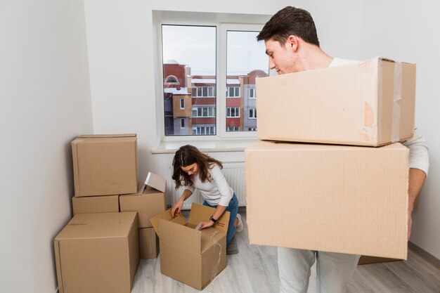 Equipaggi le scatole di cartone di trasporto che esaminano la sua amica che disimballa la scatola nella nuova casa