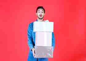 Free photo man carrying the stock of heavy silver gift boxes on red wall.
